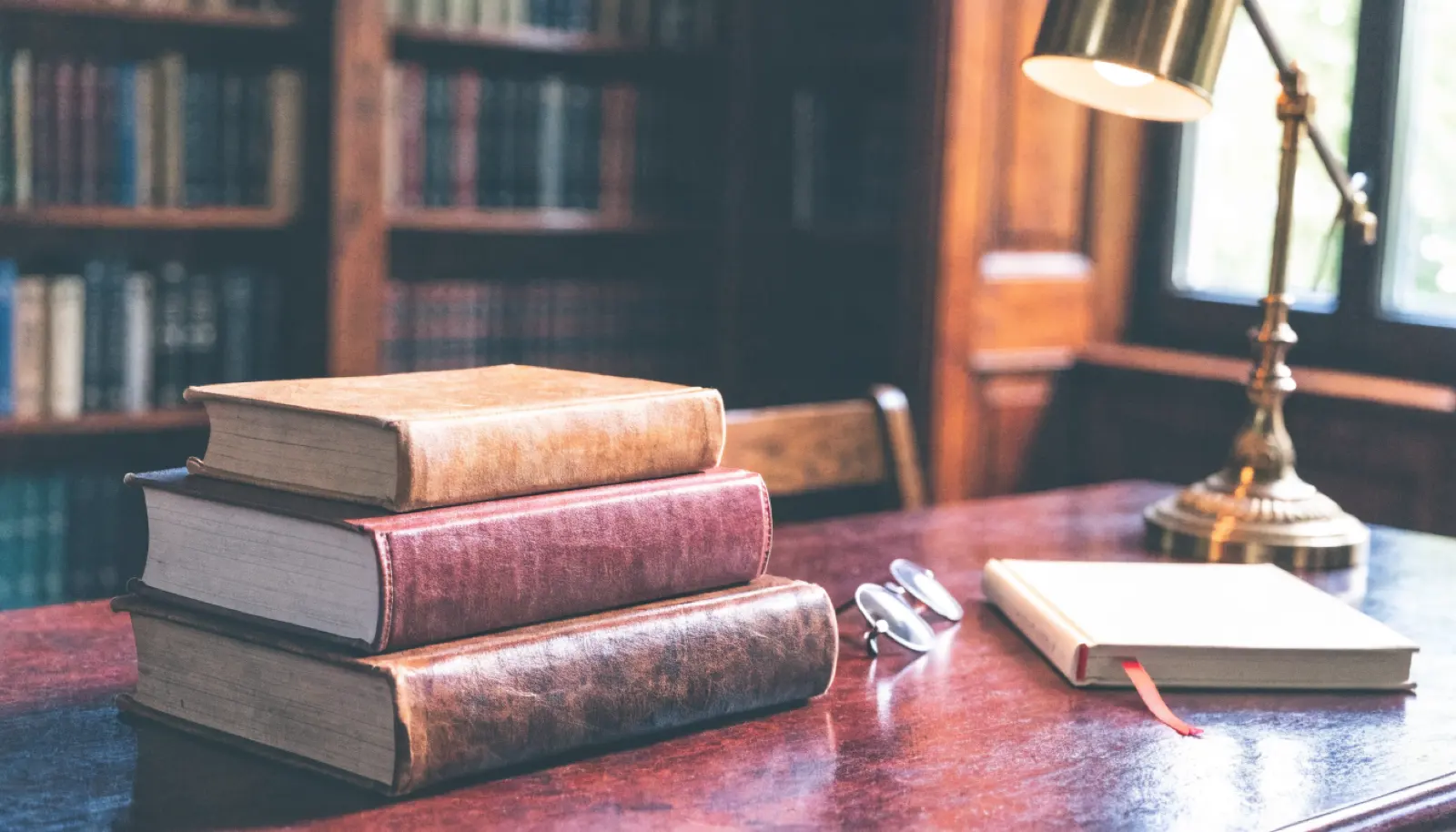 Üst üste dizilmiş psikoloji kitapları, üzerinde kapalı bir defter ve yanında yanan pirinç bir masa lambası; bordo ve arduvaz tonlarında bir çalışma masası